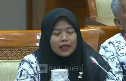 Foto tersebut merupakan tangkapan layar siaran rapat di Gedung DPR RI yang bersumber dari tayangan Parlemen/NewsTerkini. Terlihat seorang perempuan berjilbab hitam, mengenakan seragam batik, sedang menyampaikan keterangan di depan mikrofon dalam forum resmi DPR.